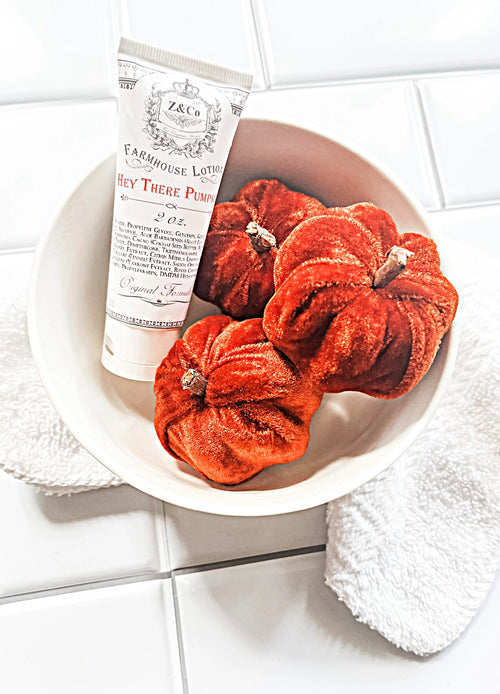 A white bowl containing three small, velvet-textured orange pumpkins and a travel-sized tube of Z&Co Hey There Pumpkin Farmhouse Lotion. The bowl rests on a white countertop with a folded white towel partially tucked underneath.