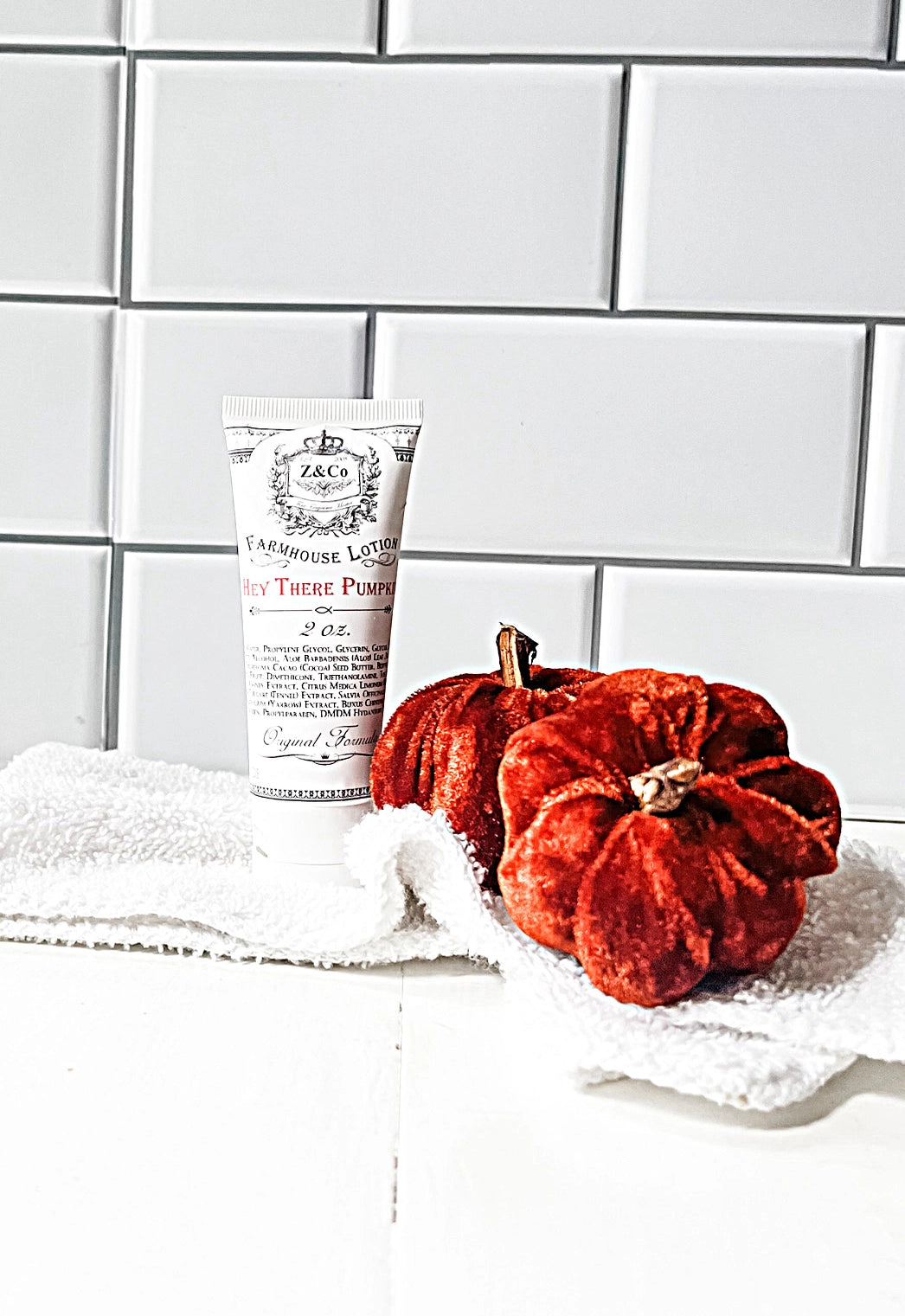 A travel-sized tube of Z&Co. Hey There Pumpkin Farmhouse Lotion rests on a white folded towel, accompanied by two small orange decorative pumpkins. The background showcases white subway tiles.