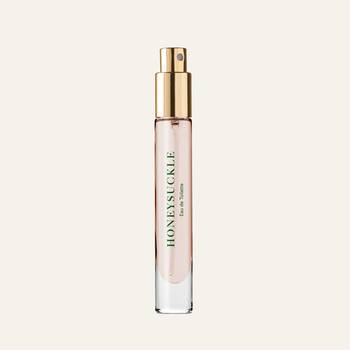 A clear glass spray bottle with a gold cap contains light pink Caswell-Massey Honeysuckle Eau de Toilette (7ml). The green label showcases this floral fragrance against an off-white background.
