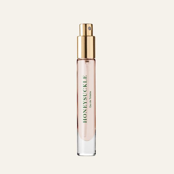 A clear glass spray bottle with a gold cap contains light pink Caswell-Massey Honeysuckle Eau de Toilette (7ml). The green label showcases this floral fragrance against an off-white background.