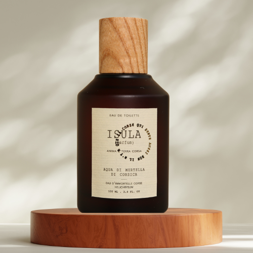 A dark glass bottle with a wooden cap rests on a wooden platform. The label reads "ISULA parfums Aqua di Murtella di Corsica - Corsican Immortelle Water eaux de toilette." Soft light creates gentle shadows nearby.