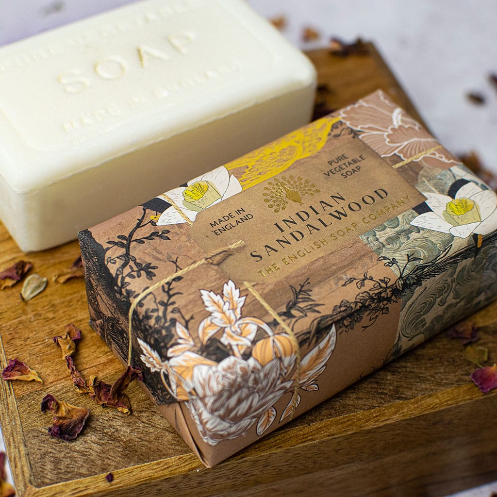 A bar of The English Soap Co. Anniversary Indian Sandalwood Soap next to its ornate box decorated with floral designs on a wooden board, surrounded by dried flowers.