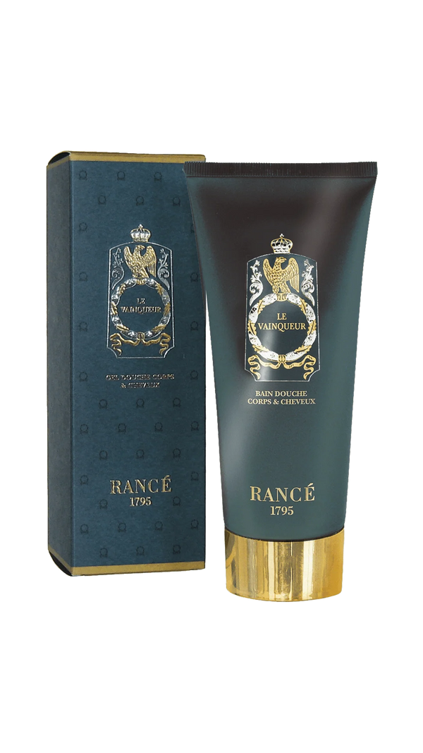A dark blue and gold tube of Rancé Le Vainqueur Body & Hair Shower Gel (200ml) stands before its matching box, both featuring an ornate gold crest and text, reflecting this men’s fragrance’s Mediterranean-inspired scent.