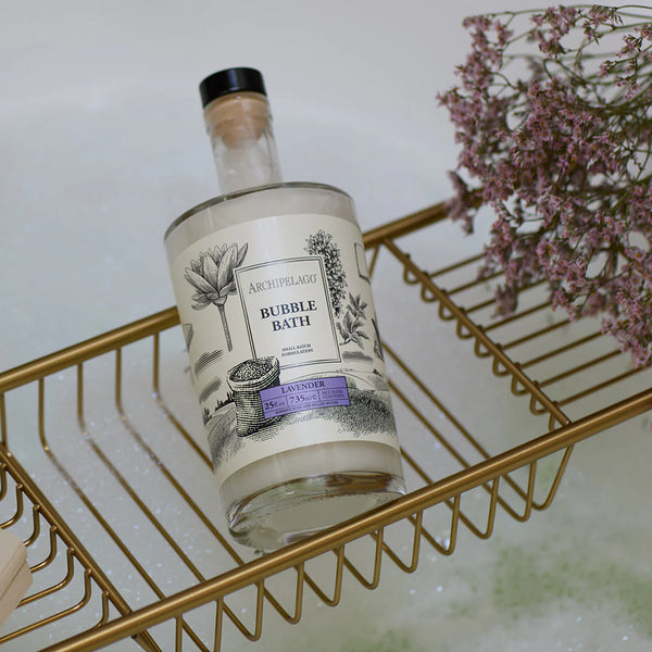 A bottle of Archipelago Botanicals Lavender 25 oz. Luxury Bubble Bath sits on a gold wire bath tray above soapy water, accented by purple flowers. The label displays a delicate botanical illustration.