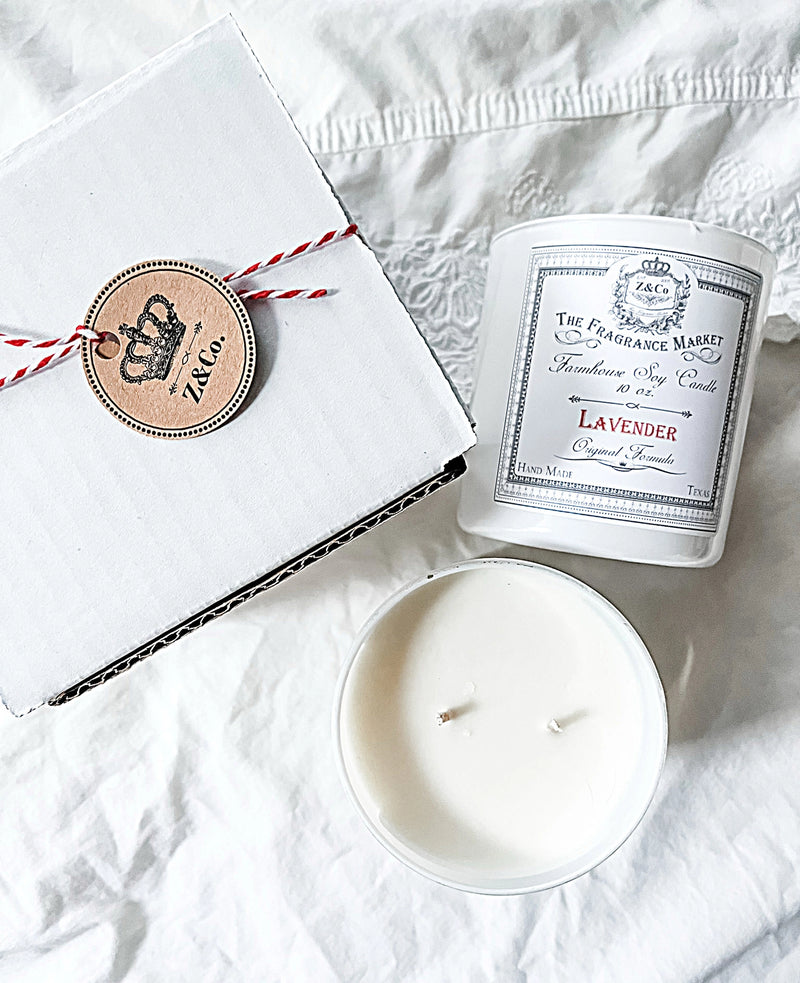 A Z&Co. Lavender Farmhouse Candle in a glass jar, with a white box and round tag, sits on soft white fabric—showcasing Z&Co.'s non-toxic, hand-crafted candles for a calming ambiance.