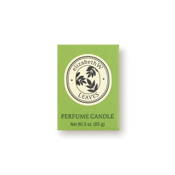 A green box labeled "elizabeth W" shows a round black leaf logo. Below: "Botanist's Scent Lab Leaves Votive Candle, Net Wt 3 oz (85 g)," a soy wax candle with Atlas cedar and sandalwood notes, sits on a plain white background.