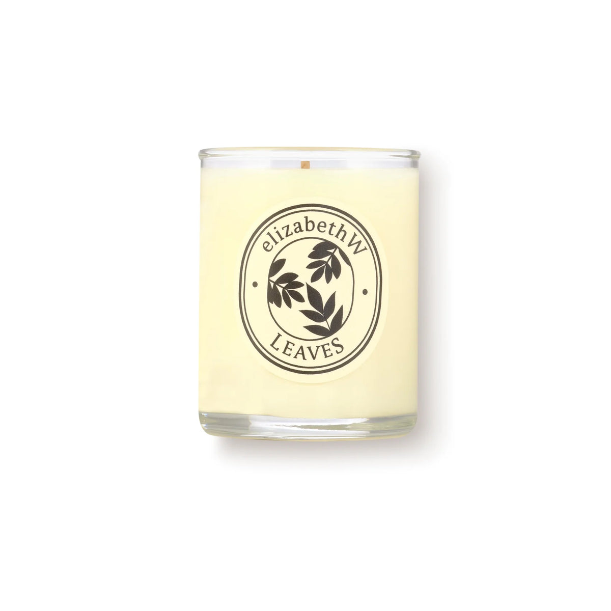 The elizabeth W Botanist's Scent Lab Leaves Votive Candle features a cream-colored soy wax in a clear glass jar with a dark leaf art label. It blends notes of Atlas cedar and sandalwood. The background is plain white.