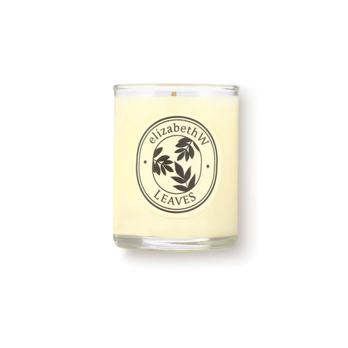 The elizabeth W Botanist's Scent Lab Leaves Votive Candle features a cream-colored soy wax in a clear glass jar with a dark leaf art label. It blends notes of Atlas cedar and sandalwood. The background is plain white.