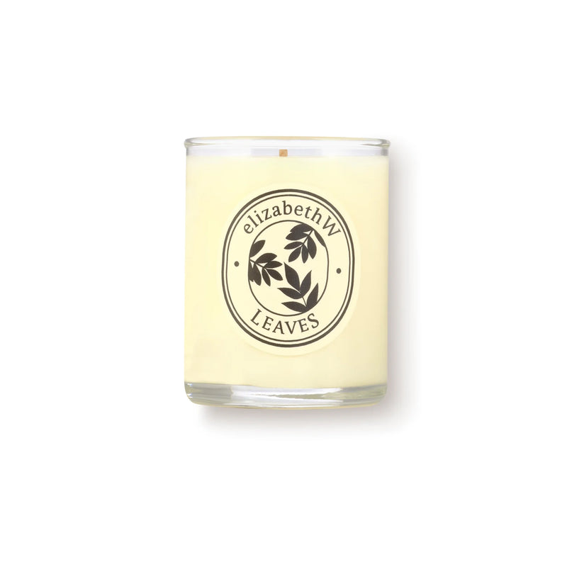 The elizabeth W Botanist's Scent Lab Leaves Votive Candle features a cream-colored soy wax in a clear glass jar with a dark leaf art label. It blends notes of Atlas cedar and sandalwood. The background is plain white.