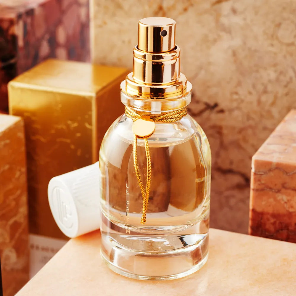 Claus Porto 1887’s Le Parfum Eau de Parfum, in a clear glass bottle with gold spray nozzle and decorative gold string, is displayed among marble-textured boxes. The white cap beside it highlights this limited edition Portuguese artisanal scent.