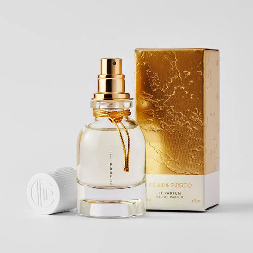 A clear glass perfume bottle with a gold spray and white cap lies next to a textured gold-and-white box labeled "Claus Porto Le Parfum Eau de Parfum" by Claus Porto 1887, a limited edition inspired by Portuguese artisanal fragrance.