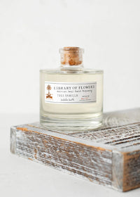 A glass bottle of Margot Elena's Library of Flowers Mini True Vanilla Bubble Bath, featuring a cork stopper and enriched with coco butter for luxury, sits on a rustic wooden surface. The clear bottle elegantly displays the light-colored liquid within.