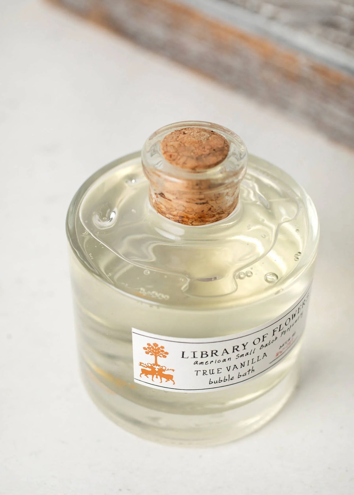 The Margot Elena "Library of Flowers" Mini True Vanilla Bubble Bath comes in a clear glass bottle with a cork stopper. It features a label with orange floral designs and "American Small Batch Perfumery," offering a luxurious experience, all resting elegantly on a light surface.