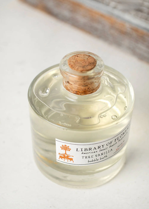 The Margot Elena "Library of Flowers True Vanilla Bubble Bath - Mini" features a subtle, warm blend of Bergamot, Dark Vanilla, and Amber in a clear glass bottle with a cork stopper. Elegantly displayed on a white surface, it holds a light-colored liquid.