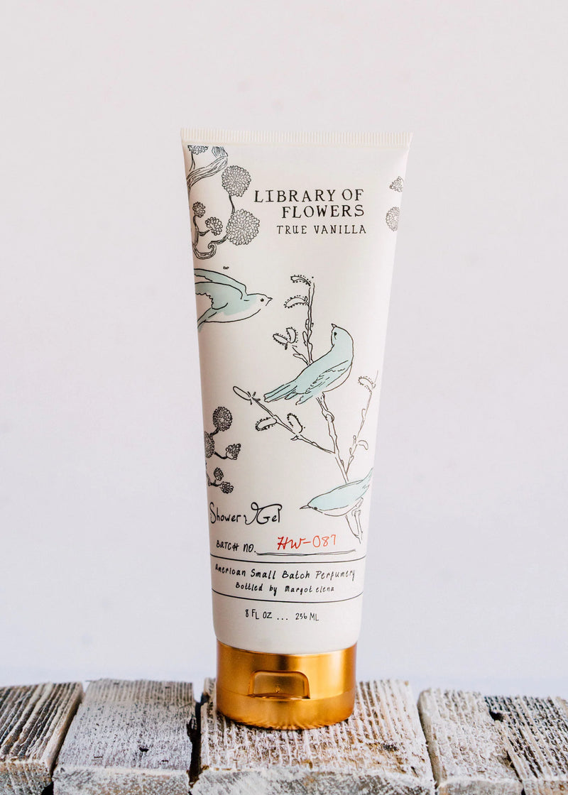 A tube of Margot Elena's Library of Flowers True Vanilla Shower Gel, enriched with Jojoba Oil, featuring an elegant floral and bird design, standing upright on a wooden surface against a white background.