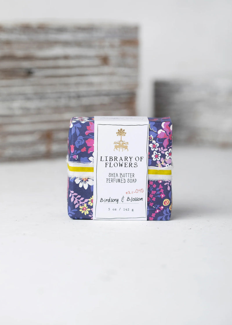 A colorful floral printed box of Margot Elena shea butter hand cream labeled as "birdsong & blossom," with notes of bergamot, positioned in front of a faintly textured white Library of Flowers Violet & Pink Roses Square Soap.