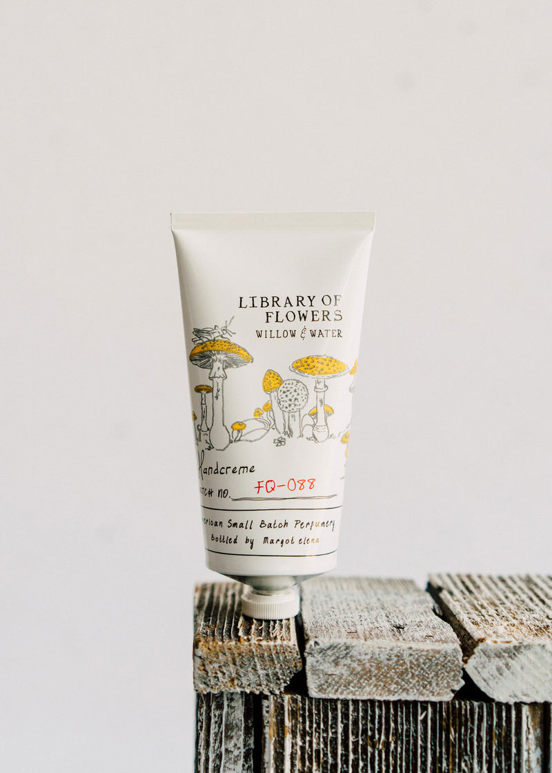 A tube of Margot Elena's Library of Flowers Willow & Water Hand Creme with shea butter rests on a stack of rustic wooden blocks, featuring whimsical illustrations and text on a white background.