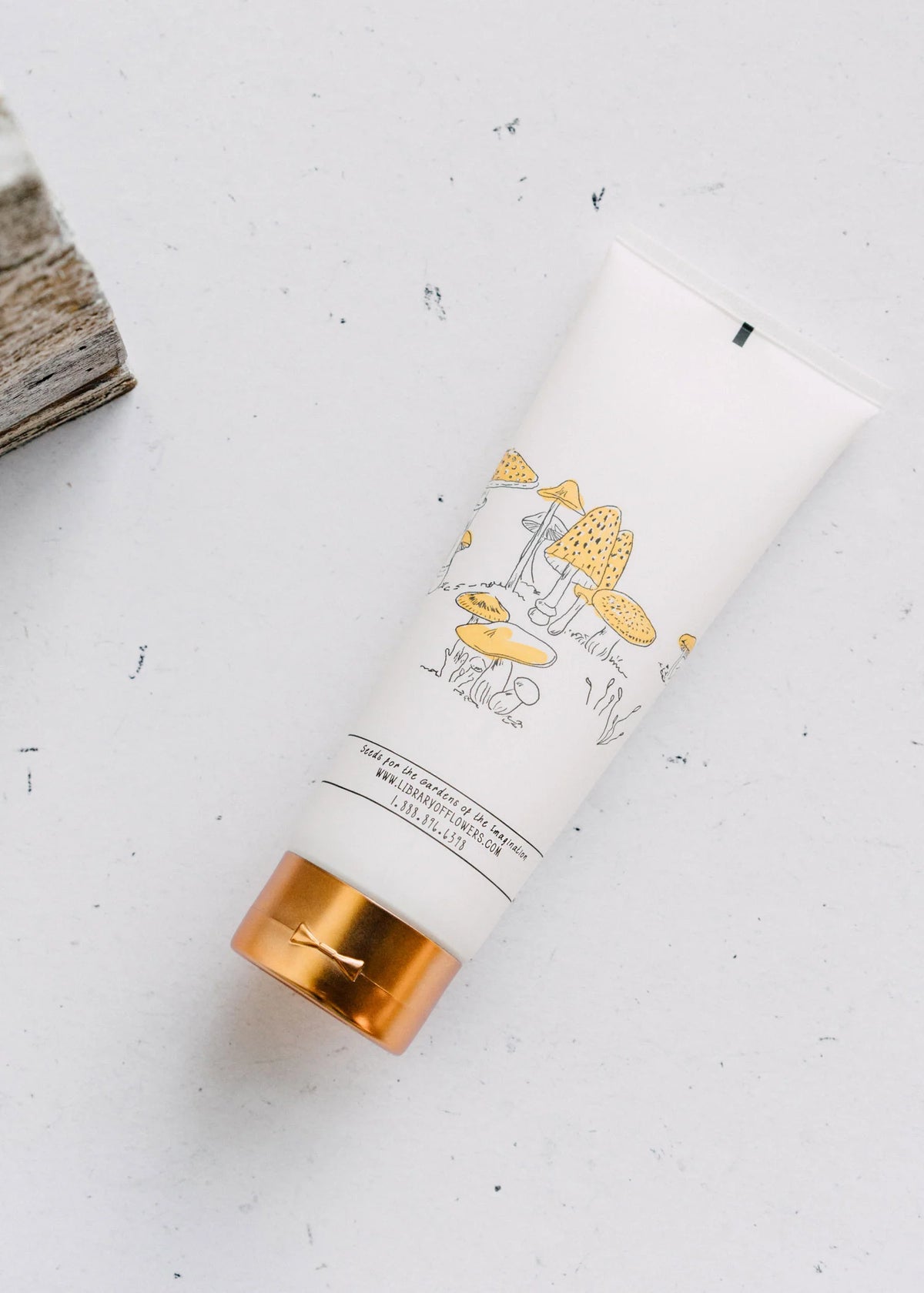 A tube of Library of Flowers Willow & Water Shower Gel with a gold cap and illustrations of mushrooms on a white background, placed next to a wooden object in the Gardens of Imagination by Margot Elena.