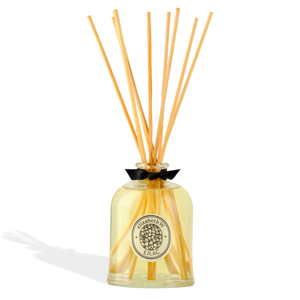 A glass bottle filled with light yellow liquid and long wooden reeds forms the Botanist's Scent Lab Lilac Diffuser by elizabeth W—a perfect home air freshener reminiscent of spring garden fragrance.