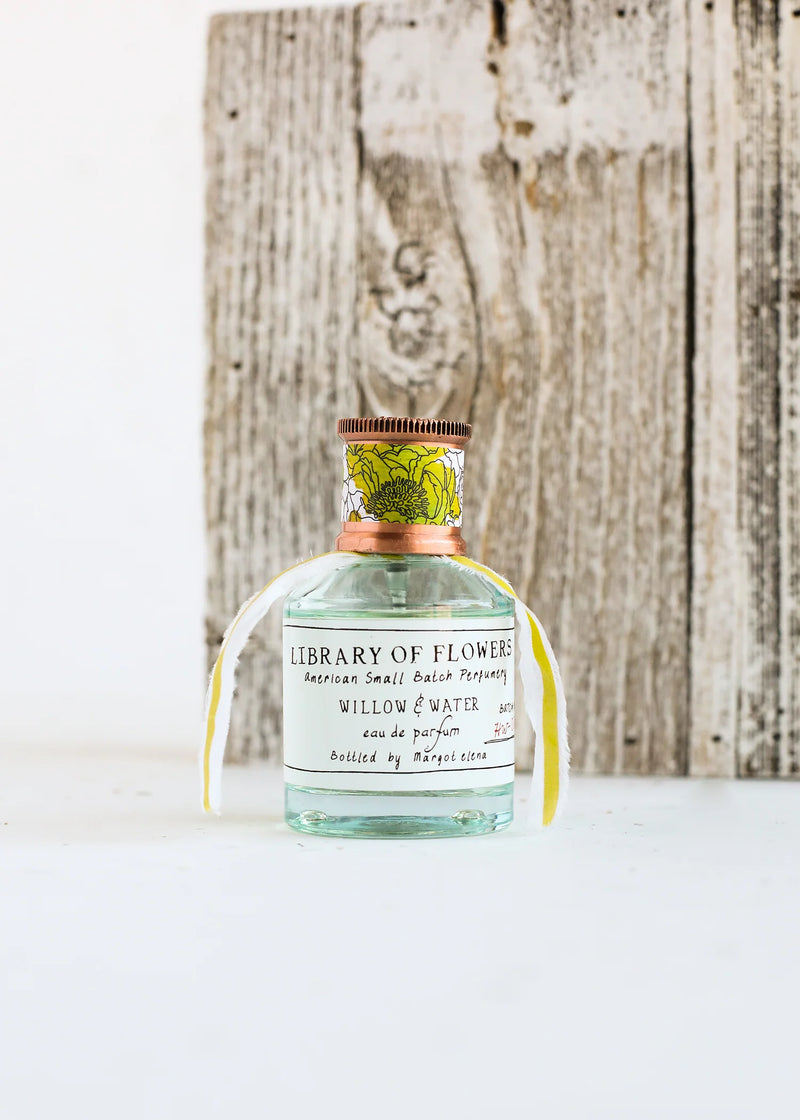 A small glass perfume bottle labeled "Margot Elena - Library of Flowers Willow & Water Eau De Parfum" stands against a rustic wooden backdrop.