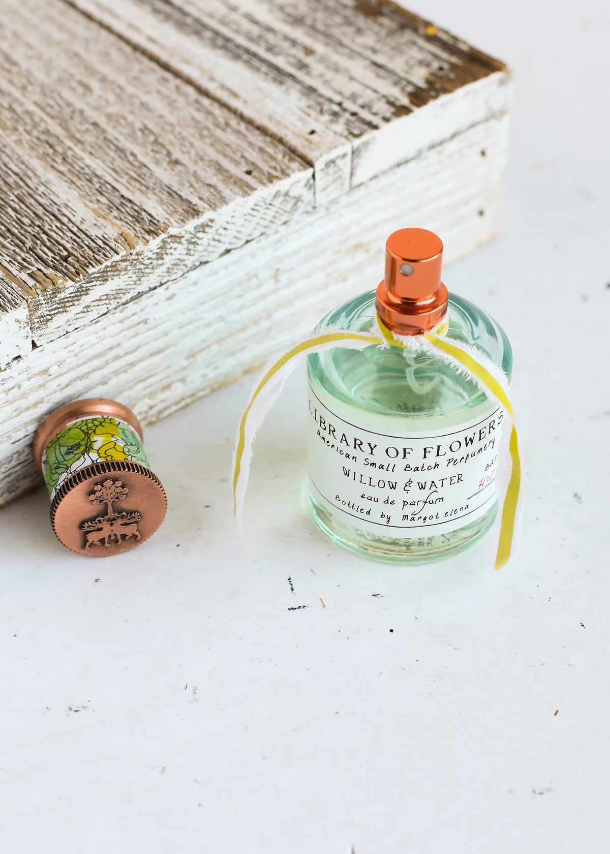 A bottle of Margot Elena's Library of Flowers Willow & Water Eau de Parfum with a floral design next to its copper-colored cap on a white surface, with a wooden box in the background.