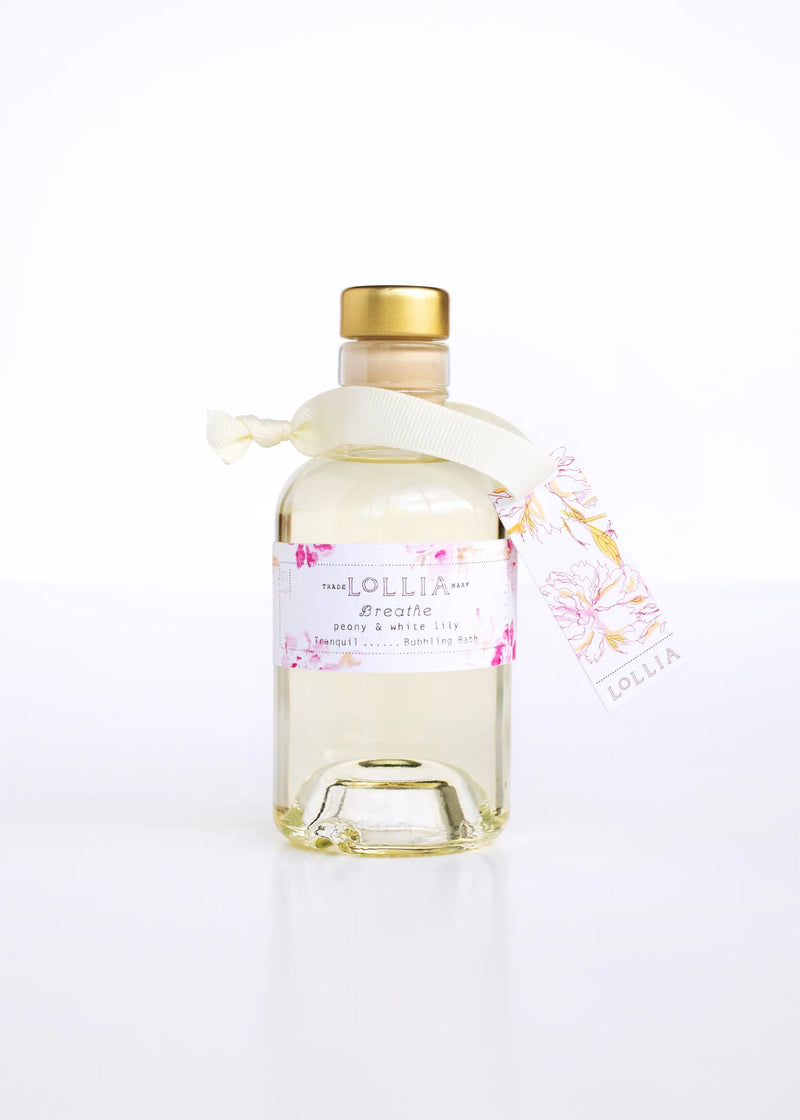 A clear glass perfume bottle with a satin ribbon and a floral tag featuring Peony & White Lily, labeled "Margot Elena" with a light golden cap, set against a white background. Product: Lollia Breathe Mini Bubble Bath