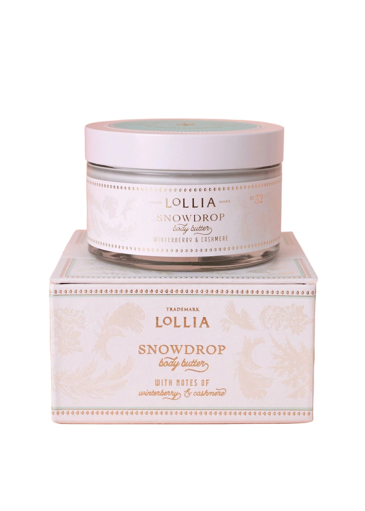 A jar of Lollia Snowdrop Body Butter by Hampton Court Essential Luxuries & The Lavender Shop rests atop its matching pastel box with white and gold floral designs, set against a pale pink background.