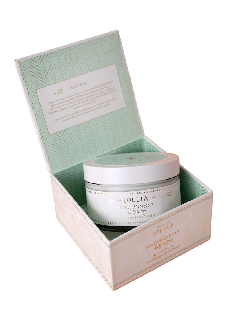An open pastel green and white box reveals a round jar of Lollia Snowdrop Body Butter by Hampton Court Essential Luxuries & The Lavender Shop, labeled No. 32, on a pale pink background. The inner lid features a printed description.