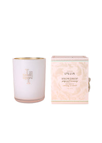 The Margot Elena Lollia Snowdrop Luminary features a white candle in a glass holder with gold rim and emblem, beside a pink and gold patterned box. Made with a luxurious soy and coconut wax blend.
