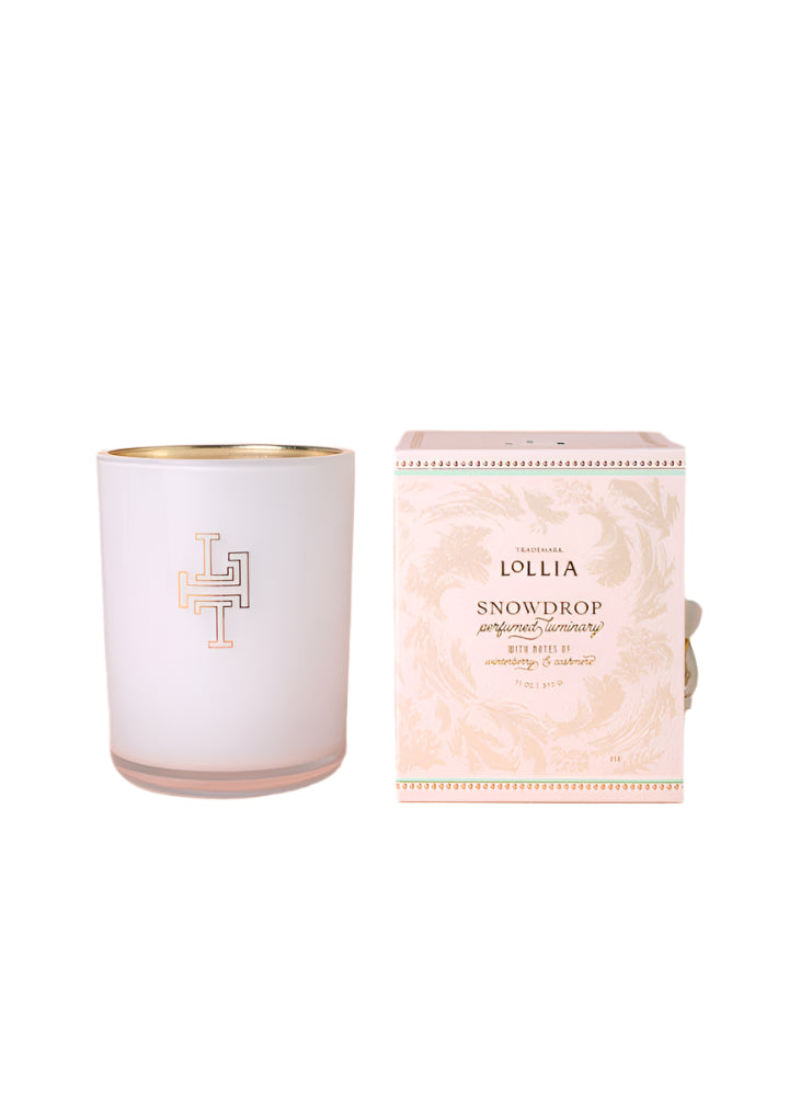 The Margot Elena Lollia Snowdrop Luminary features a white candle in a glass holder with gold rim and emblem, beside a pink and gold patterned box. Made with a luxurious soy and coconut wax blend.