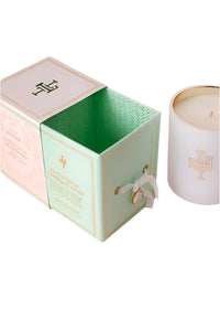 The Margot Elena Lollia Snowdrop Luminary candle, made from a luxurious soy and coconut wax blend, sits in a glass holder with a gold emblem beside its open mint green and pink decorative box with gold details and a white ribbon pull.