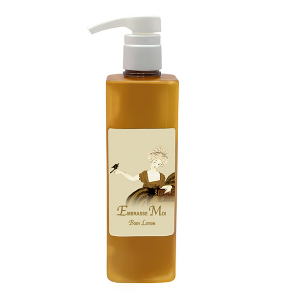 A rectangular amber bottle with a white pump labeled "La Bouquetiere Embrasse Moi Body Lotion" features vintage art of a woman holding a bird. Enriched with African Shea Butter and soothing Aloe Vera by La Bouquetiere.