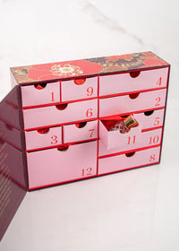 A pink and red Margot Elena Advent Calendar with gold detailing and numbered drawers from 1 to 12. One drawer, numbered 11, is partially open, revealing a small wrapped gift inside. The box is set on a white marble surface.