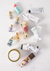 An assortment of beauty and wellness products displayed on a white surface. Items include the Margot Elena Advent Calendar, featuring small bottles of perfume, tubes of hand cream, tins of lip balm and travel-sized containers of lotions. The packaging features floral and elegant designs from Margot Elena.
