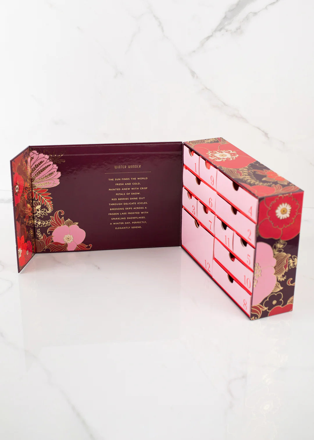 A Margot Elena Advent Calendar with floral and ornate designs, containing twelve small drawers numbered 1 through 12. The box is open, revealing a message inside the lid. The background is a white marble surface.