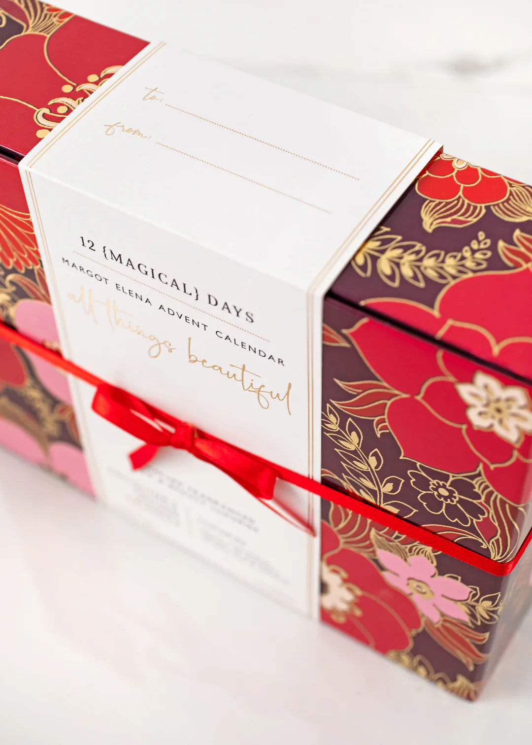 A festive Margot Elena Advent Calendar is decorated with floral patterns in red and gold. It is labeled "12 Magical Days" and wrapped with a red ribbon. There is a space for writing "To" and "From" on the top. The text "all things beautiful" is elegantly written on it.