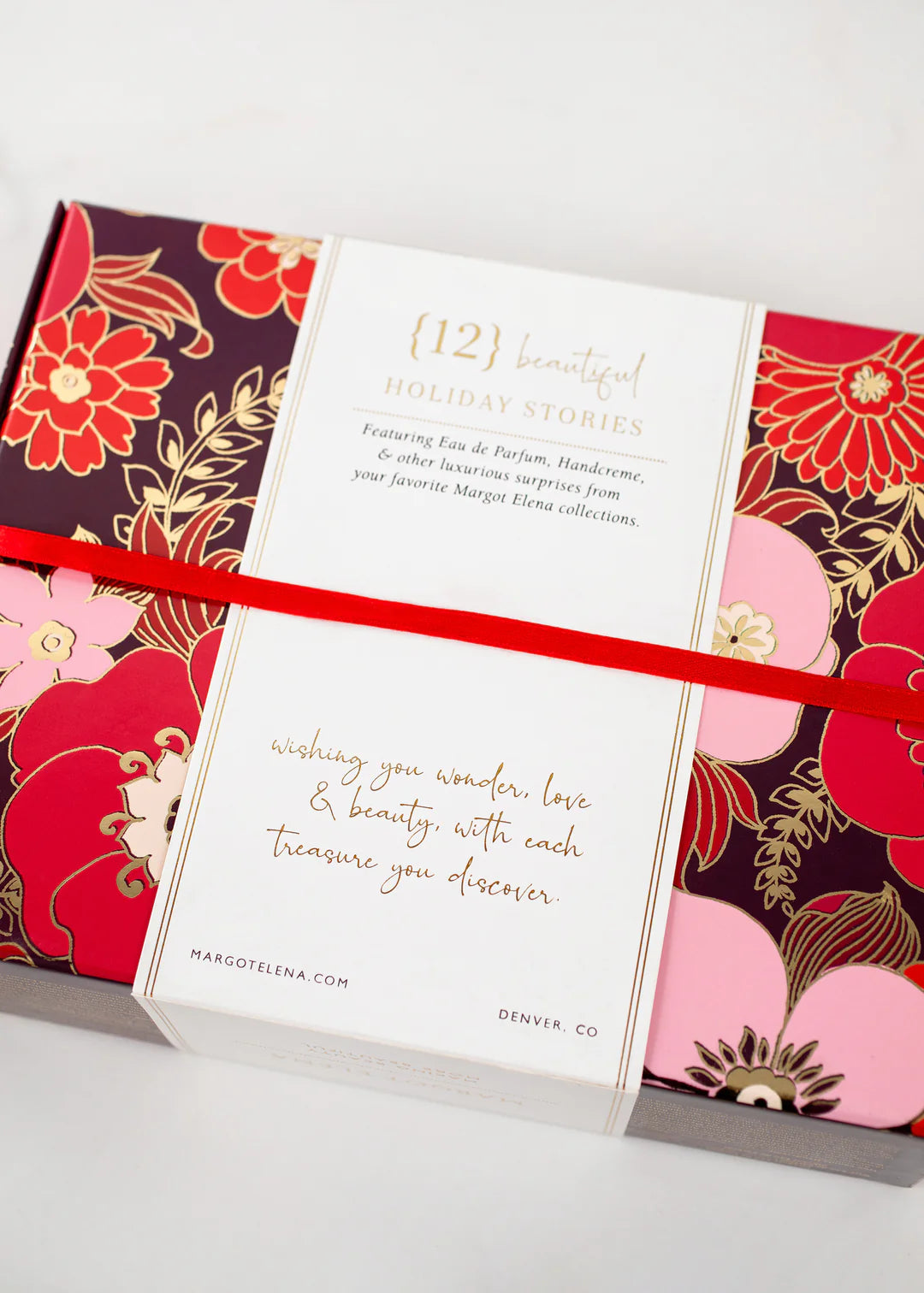 A decorative box adorned with floral designs in red, pink, and gold. A white band with text in elegant gold and red font wraps around the box, reading: "Margot Elena Advent Calendar" by Margot Elena and "wishing you wonder, love & beauty with each treasure you discover.