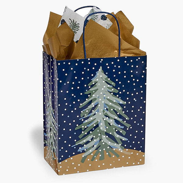 The Nashville Wraps Midnight Flurry Paper Gift Bag, 8x4.75x10", features a snow-covered pine on a dark blue background with white dots—ideal for holiday gifts and fully recyclable. Includes brown and pine-printed white tissue.