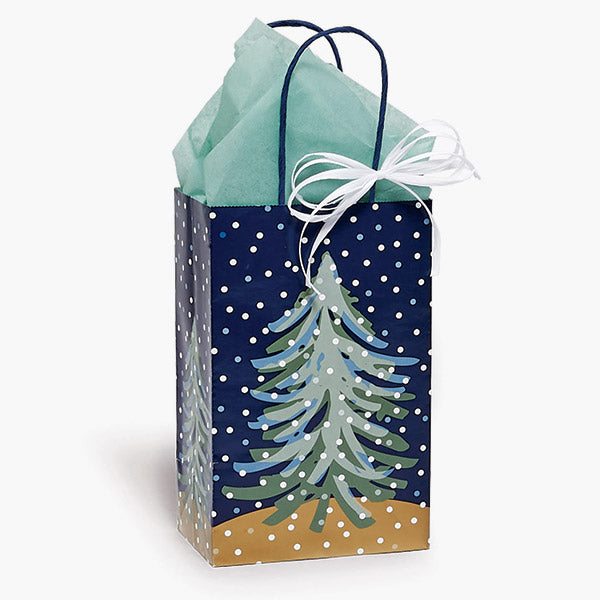 The Nashville Wraps Midnight Flurry Paper Gift Bag (5.25x3.50x8.25") features a snowy evergreen design on a blue background with white dots, green tissue paper, and a white ribbon—an eco-friendly recycled choice for gifting.