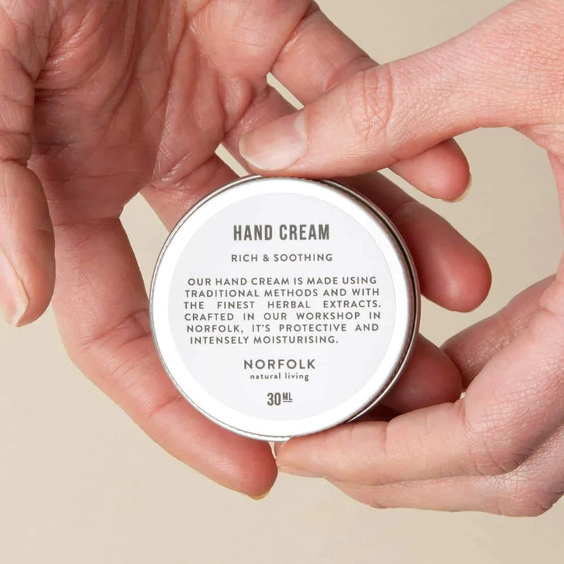 Two hands hold a small container of Norfolk Natural Living Midsummer 24 Nourishing Hand Cream labeled as rich and soothing, made with shea butter. The text emphasizes its moisturizing and protective qualities.