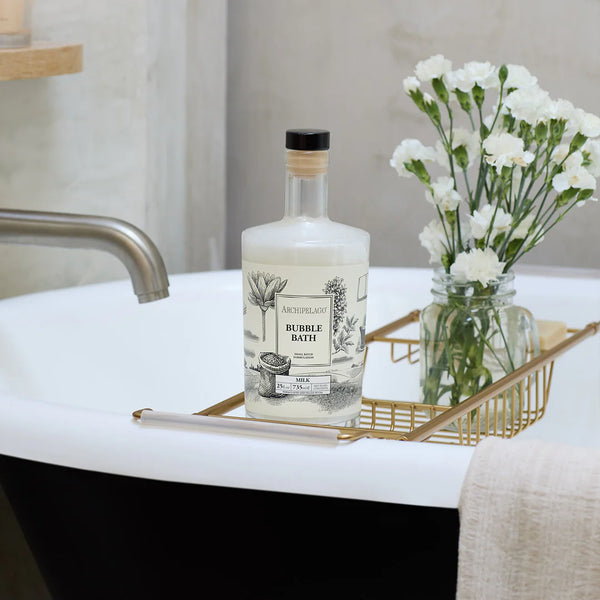 A bottle of Archipelago Botanicals Milk 25 oz. Luxury Bubble Bath, featured on Oprah's Favorite Things 2024, rests on a gold tray atop a white tub next to white flowers and a folded towel, creating a soothing, elegant bathroom scene.