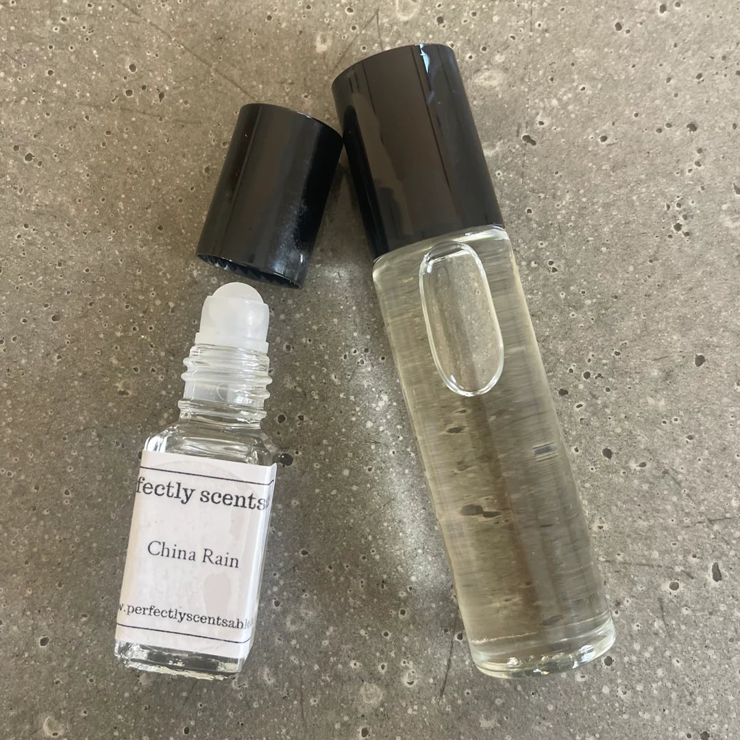 Two clear glass perfume bottles with black caps from Perfectly Scentsable, including a China Lily Mini Rollerball lying open and a taller bottle standing upright, both on a textured gray surface.