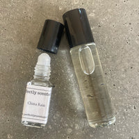 Two clear glass perfume bottles with black caps from Perfectly Scentsable, including a China Lily Mini Rollerball lying open and a taller bottle standing upright, both on a textured gray surface.
