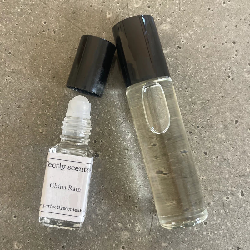 Two clear glass perfume bottles with black caps from Perfectly Scentsable, including a China Lily Mini Rollerball lying open and a taller bottle standing upright, both on a textured gray surface.