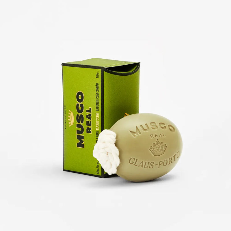 A bar of Claus Porto Musgo Real Classic Scent Soap on a Rope next to its green packaging on a white background. The soap is oval-shaped with embossed text and a decorative white element.