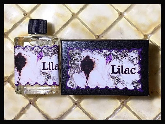 A small bottle and box, labeled "Seventh Muse Fragrant Oil - Lilac" from Seventh Muse Fragrant Oils, sit on a light-textured surface. The labels feature floral designs with purple accents, evoking a vintage French perfume vibe.