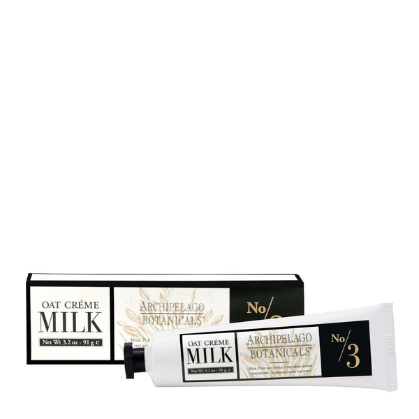 A white tube of Archipelago Oat Milk Hand Crème by Archipelago Botanicals rests in front of a matching box with gold accents and botanical designs, highlighting its luxurious, soothing formula with jojoba esters.