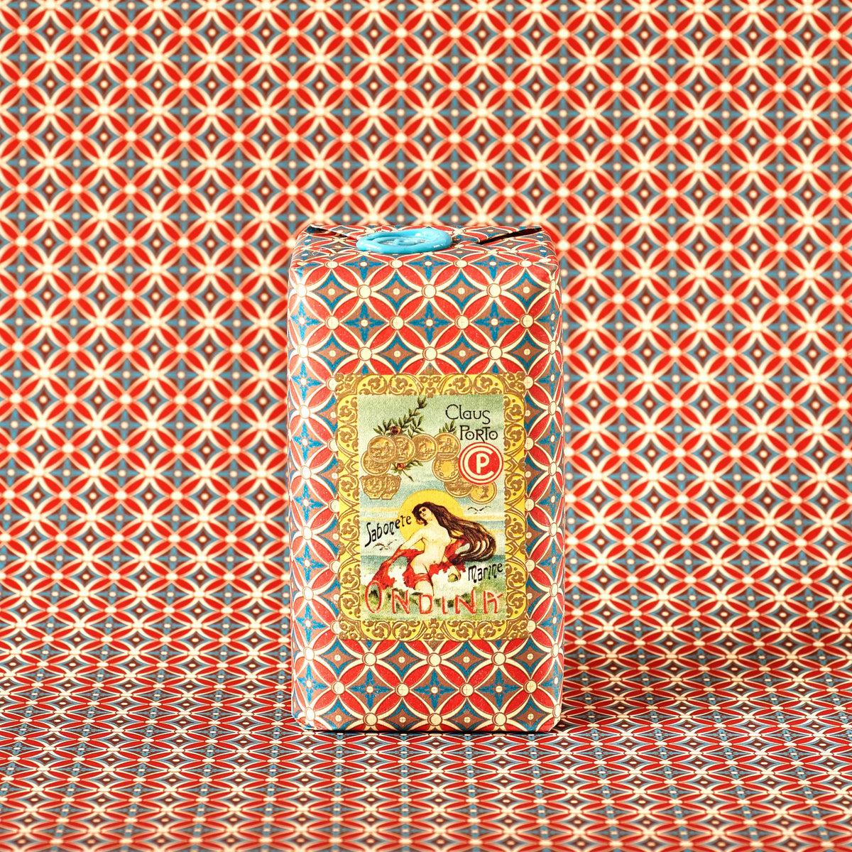 A rectangular box with red, blue, and gold geometric patterns blends into a matching background. An ornate vintage label features a woman and the text "Claus Porto 1887 Classico Odina Sea Mist Soap," enriched with mango seed oil.