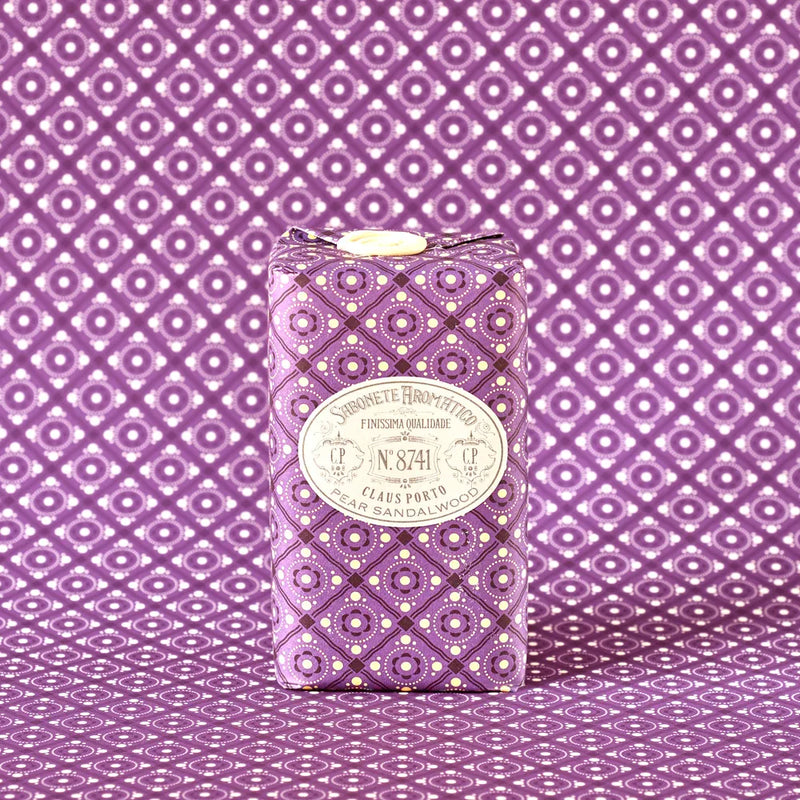 A bar of Claus Porto 1887 Classico 8741 Pear Sandalwood Soap, wrapped in purple patterned paper with white accents, rests on a matching surface, suggesting a fruity-woody scent and nourishing pistachio nut seed oil.
