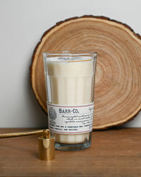 A Barr-Co. Original Scent Parfait Candle sits on a wooden surface with a tree slice in the background and a gold snuffer nearby, offering a long-lasting burn in the comforting Barr-Co. signature fragrance.